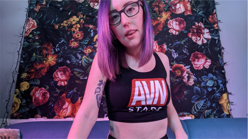 Tattooed femdom AVN Stars model Miss Faith Rae poses seductively in her AVN Stars branded crop top and high waisted shorts while looking down at the viewer.